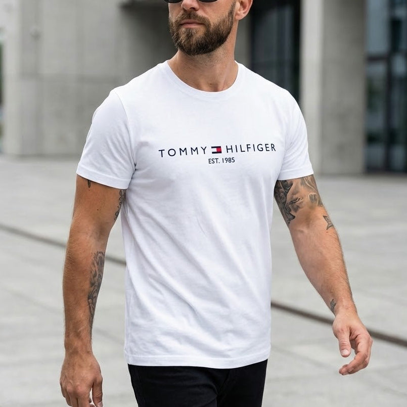 Мъж със слънчеви очила и татуировки в бяла тениска Tommy Hilfiger с лого и надпис Est. 1985